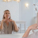 Beautiful senior woman looking at herself at mirror doing face fitness - self face massage, sitting in front of mirror at home, making beauty routine procedures